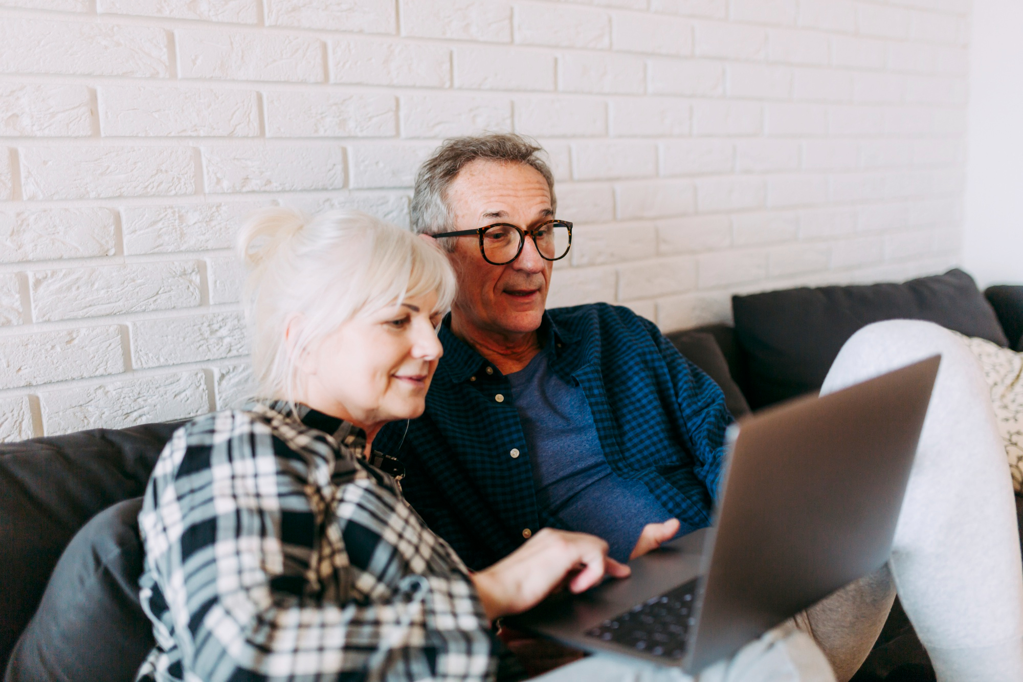 Uśmiechnięta para seniorów na kanapie wspólnie korzysta z laptopa, przeglądając treści w internecie.